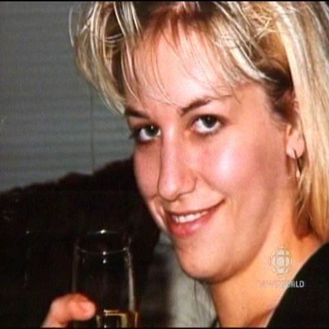 Karla Homolka is smiling, while holding a champagne glass, at the back is a white blinds, has middle-length blonde hair, and is wearing a black shirt.