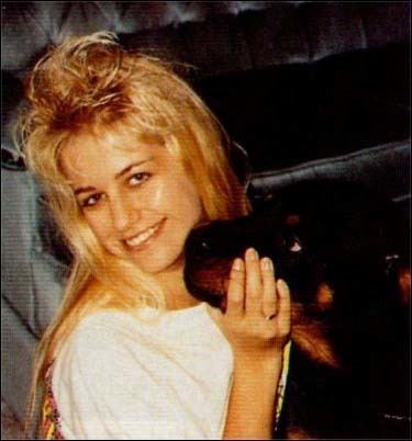 Karla Homolka is smiling, sitting on the floor in front of the black sofa, while holding her black dog’s mouth with her right hand, has long blonde hair wearing a gold ring and a white shirt.