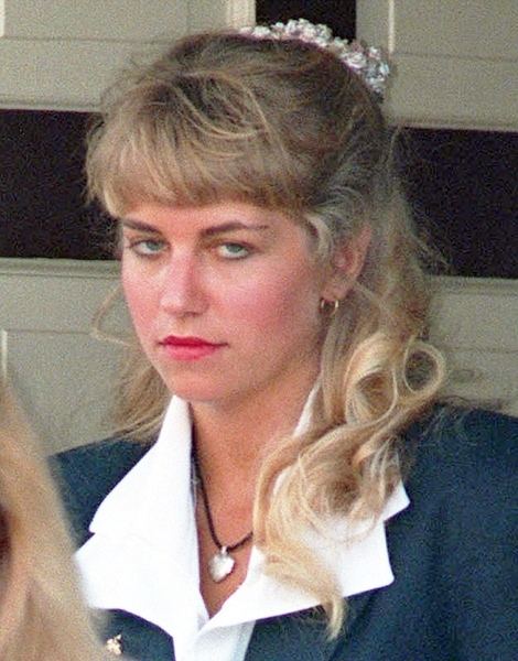 Karla Homolka is mad, leaning backwards in a wall with black line, has long blonde hair white tie, wearing gold earrings, necklace with silver heart pendant and white top under a blue-green coat.