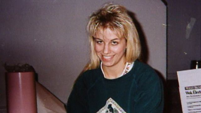 Karla Homolka is happy, sitting in a room with an orange tube at her right and gray cabinet behind her left, has middle-length blonde hair, wearing a blue-green printed shirt.