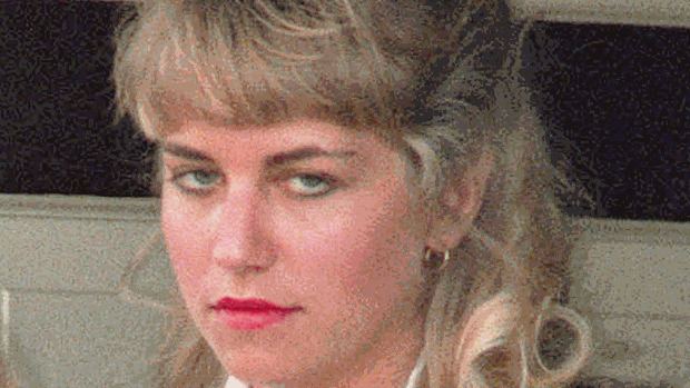 Karla Homolka is mad, leaning backwards in a wall with black line, has blonde hair, wearing gold earrings, and white top.