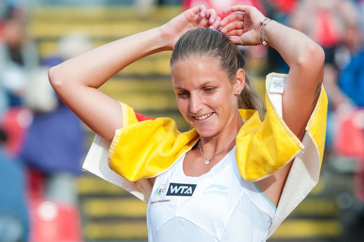 Karolína Plíšková smiling, wearing a white sleeveless top with a yellow towel on her shoulder while holding her hair.