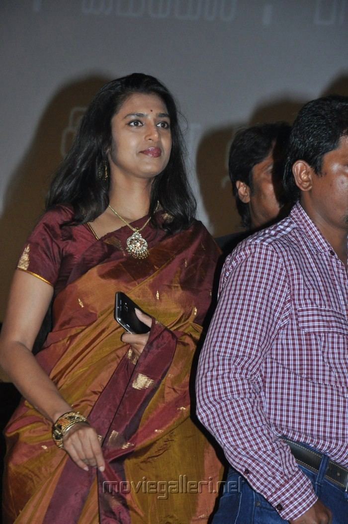 Kasthuri looking afar while holding a cellphone and wearing a brown and maroon dress and some pieces of jewelry