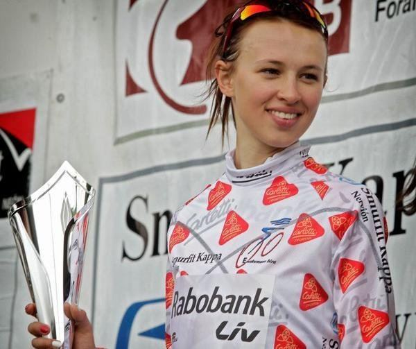 Katarzyna Niewiadoma smiling while holding a trophy and wearing a white and orange long sleeve jersey and sunglasses on her head