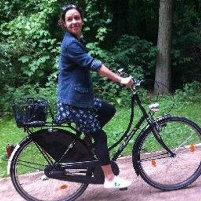 Kate Atkinson riding on a bicycle while wearing a denim jacket, black leggings, and white shoes