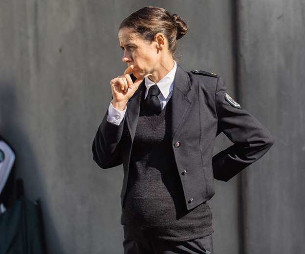 Kate Atkinson's hand on her lips while wearing a black coat, black vest, necktie, and white long sleeves