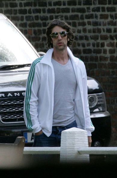 Richard Ashcroft wearing sunglasses, a white jacket, a gray shirt, and blue jeans.