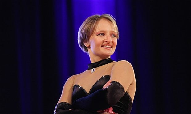 Katerina Tikhonova is smiling with arms crossed on her chest, with short blonde hair, and wearing a black leotard with a diamond.