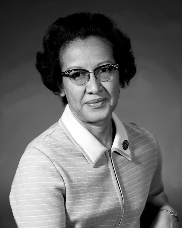 Katherine Johnson with a tight-lipped smile, black short hair, wearing eyeglasses, a watch, and a striped top with a white collar with a pin.