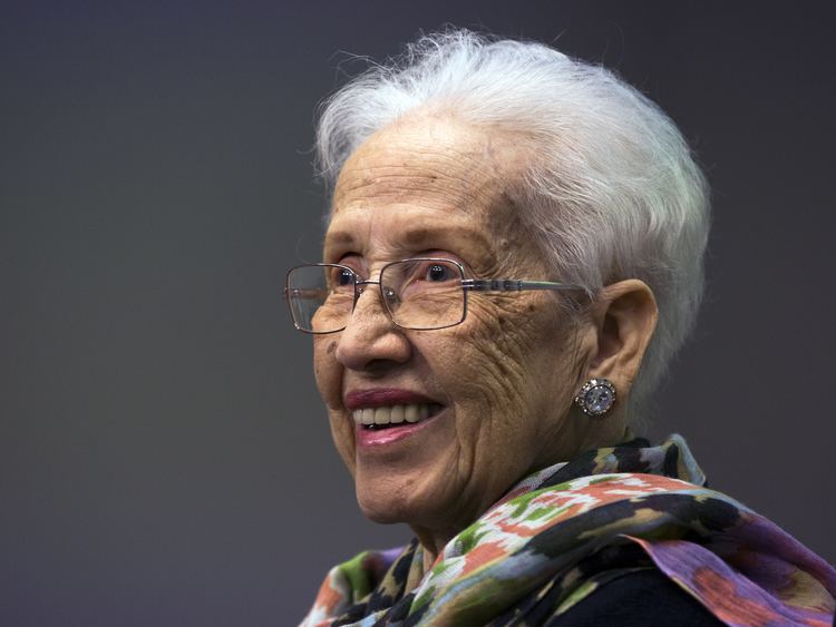 Katherine Johnson with a big smile on her face, short white hair, wearing eyeglasses, earrings, a black top, and a multi-colored scarf.