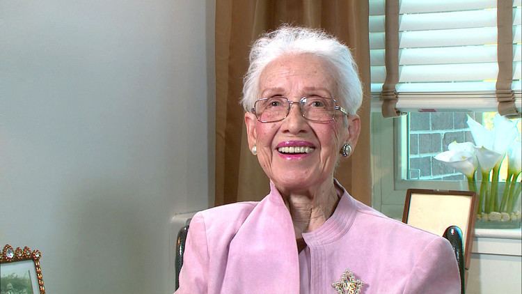 Katherine Johnson smiling while sitting on a steel chair, with short white hair, wearing eyeglasses, earrings, and a pink blazer with a golden pin.