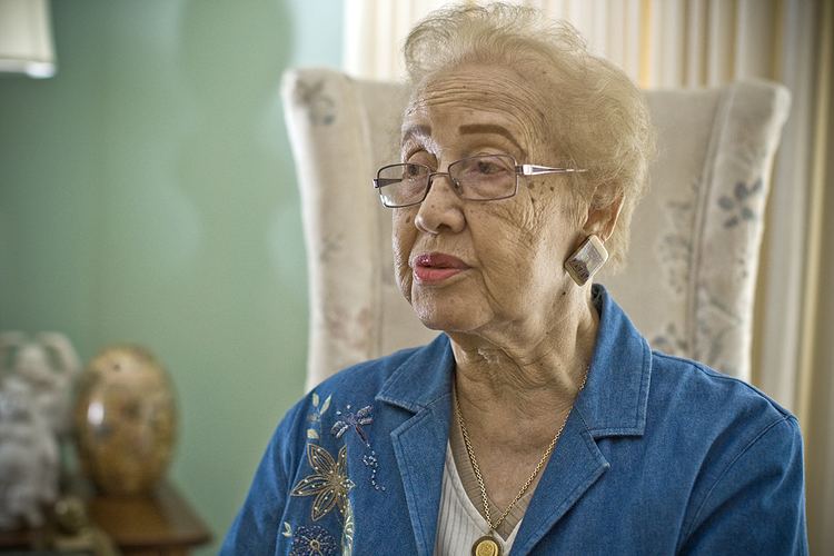 Katherine Johnson with a serious face while pouting her lips and sitting on a white chair, with short blonde hair, wearing eyeglasses, earrings, a necklace, and a denim jacket with flower design over a white top.