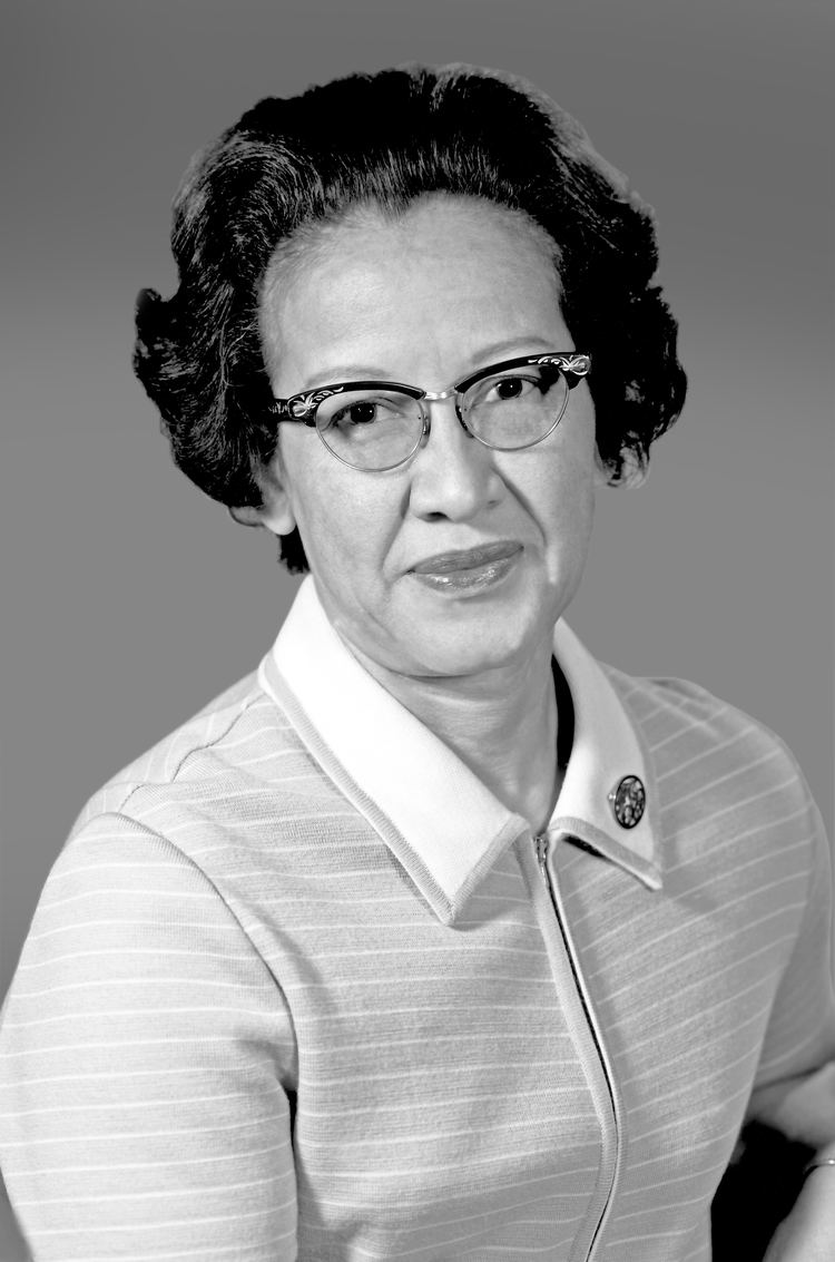 Katherine Johnson with a tight-lipped smile, black short hair, wearing eyeglasses, a watch, and a striped top with a white collar with a pin.
