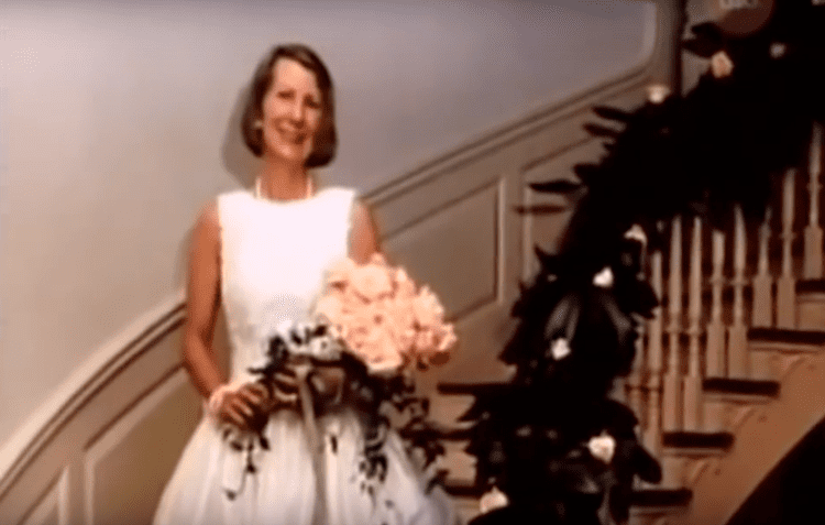Kathleen Peterson smiling and holding a bouquet of pink flowers with green leaves while standing on a staircase with green leaves on its handrail. She has a blonde short hair, wearing a pearl earring, pearl necklace, a white bracelet, and a white sleeveless gown