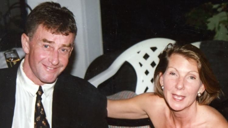 Kathleen Peterson (right) smiling with her arms on Michael’s (left) back while holding a glass of wine and Michael is smiling with a white chair in their background. Kathleen has a blonde short hair, wearing a pearl earring while Michael has a black hair wearing a white polo with a black necktie under a black coat