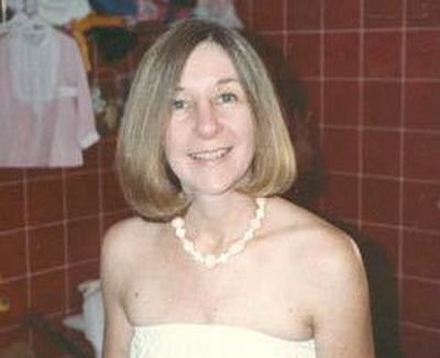 Kathleen Peterson smiling with hanged clothes and red tiles in her background, she has a blonde short hair wearing a white necklace and a white tube