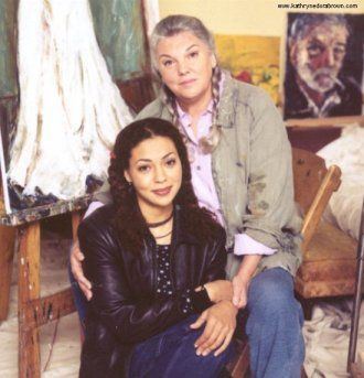 Kathryne Dora Brown smiling and sitting beside her mother Tyne Daly with paintings in the background. Kathryne with curly hair is wearing a black necklace and a black blouse under a black leather jacket and jeans while Tyne with gray braided hair is wearing a pink polo shirt under a brown jacket and jeans