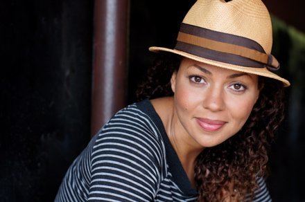 Kathryne Dora Brown smiling with curly hair and a brown pole in the background, wearing  a brown hat and blouse with black and white stripes