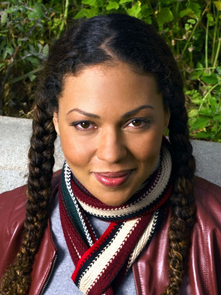 Kathryne Dora Brown smiling with braided hair and plants in the background, wearing a knitted scarf, and a gray blouse under a red leather jacket