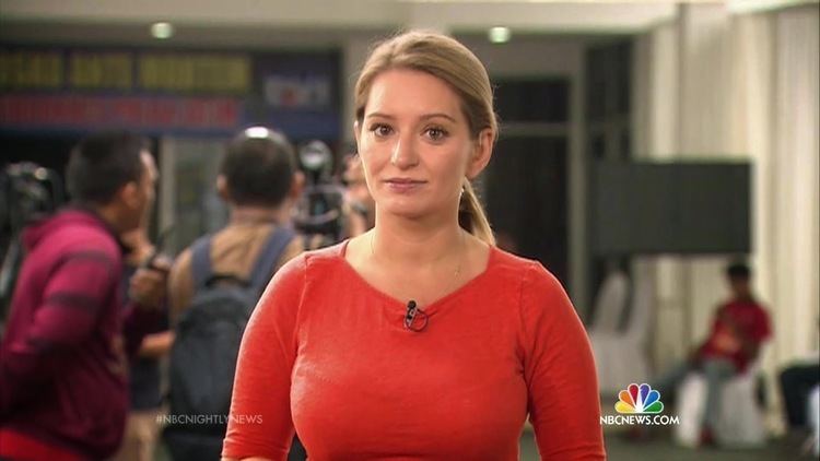 Katy Tur smiling with people in her background in a photo captured from NBC news, she has tie blonde hair, wearing a necklace, and a lav mic on  a red long sleeve