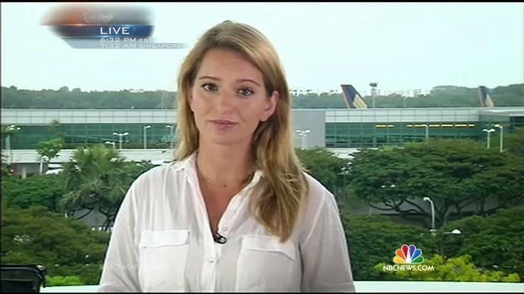 Katy Tur smiling with trees and a building in her background in a photo captured from NBC news, she has blonde hair, wearing a necklace, a lav mic on  a white polo longsleeve