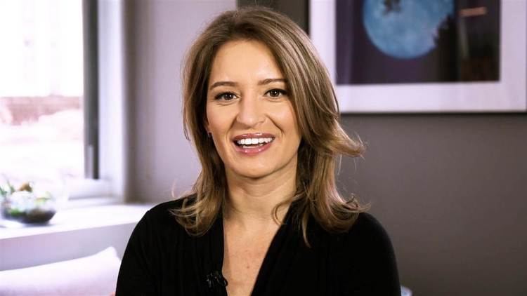 Katy Tur smiling inside of an office, she has blonde hair, wearing a lav mic on  a  black long sleeve