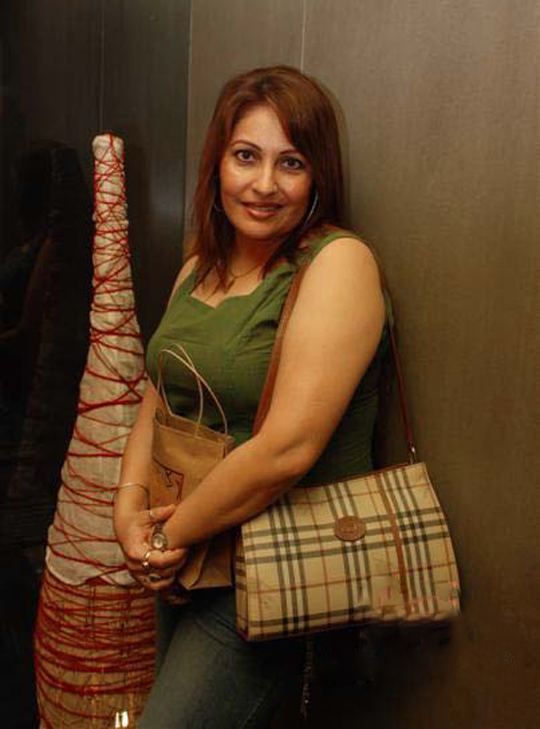 Kavita Kapoor smiling and leaning on a wall and carrying a paper bag while wearing a green sleeveless shirt with a handbag on her shoulder.