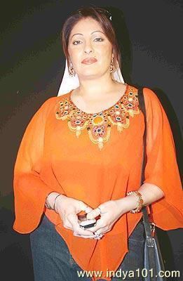 Kavita Kapoor posing and holding her phone while wearing an orange blouse and jean pants along with various accessories.