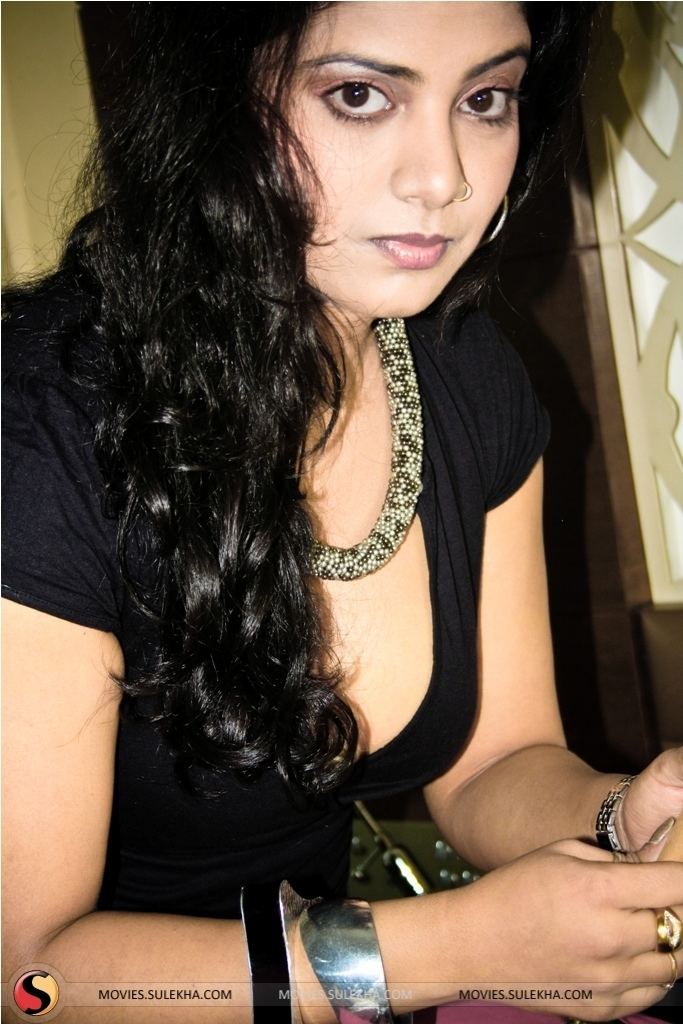 Kavita Radheshyam is serious, has black hair and a nose piercing on her left wing of the nose, wears a wristwatch in her left hand, a silver and black bracelet, and rings on her right hand, a white-green small beads necklace, and a black cleavage showing top.