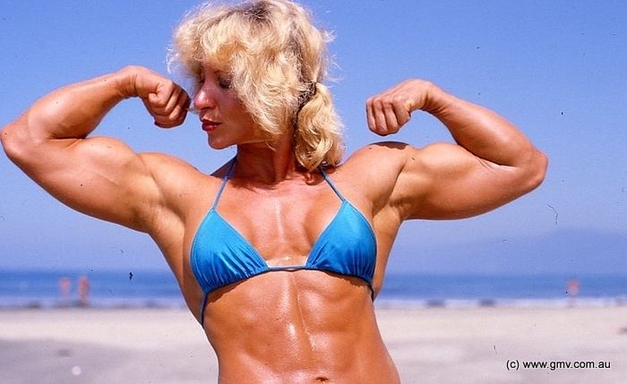 Kay Baxter doing a "Front Double Biceps" pose at the beach while wearing a blue top