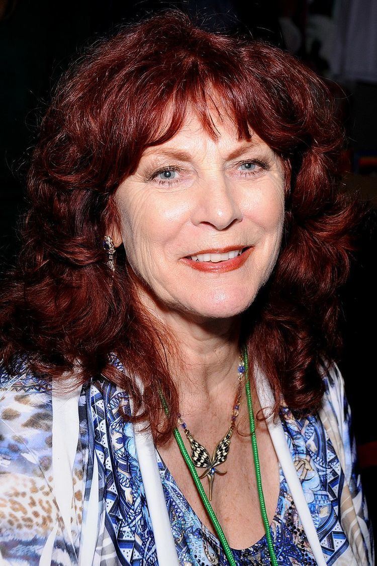 Kay Tylor Parker smiles while wearing a blue printed shirt, a necklace, and pair of earrings