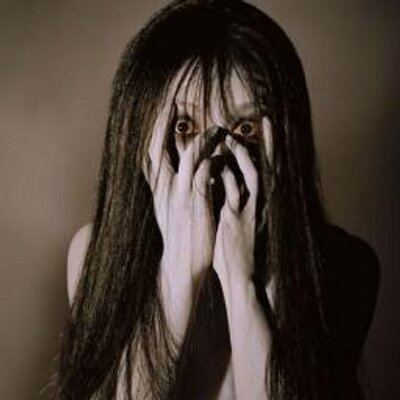Kayako Saeki is serious, has a frightening expression in brown background, eyes wide open, hands on her face covering the mouth and nose, has long black hair and white pale skin.
