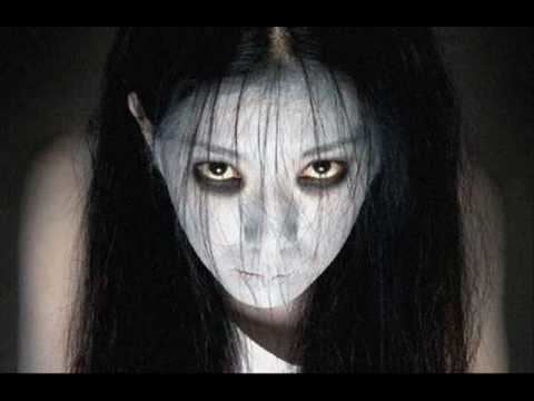 Kayako Saeki is serious, has a frightening expression in black background, eyes wide open, has long black hair and white pale skin wearing a white top.
