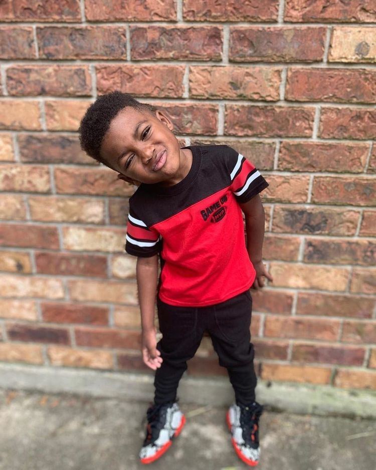 Kayden Gaulden pouting his lips while leaning on the wall, with curly hair, wearing a white, black and red shirt and shoes, and black pants.