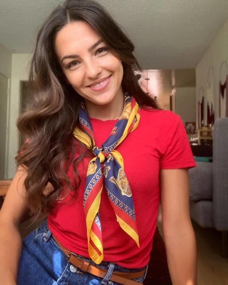 Kayla Compton smiling, with wavy long hair, wearing a colorful ribbon scarf, red shirt, and blue jeans with a brown belt.
