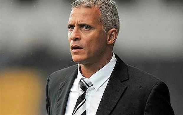 Keith Curle is serious, mouth half open, has gray hair, wearing a white polo and white necktie with black pattern under a black suit.