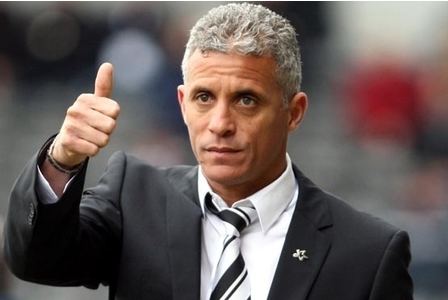 Keith Curle is serious, standing giving thumbs up with his right hand, he has gray hair, wearing a black strap watch, a white polo along and a white necktie with black pattern under a black suit with silver pin on its chest.