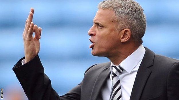 Keith Curle is serious, facing to his right, mouth half open, giving a sign with his three fingers, he has gray hair wearing, a white polo and white necktie with black pattern under a black suit.