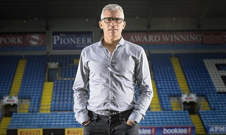 In a stadium with yellow layered steps and blue chairs, with different advertisement banner “PORK in red banner, PIONEER in blue banner and award winning in red banner” at the back,  Keith Curle is serious, standing in the middle of the stadium, both hands in his pocket, he has white hair, wearing a silver watch a gray long sleeve polo, a black eyeglass and a black denim pants.