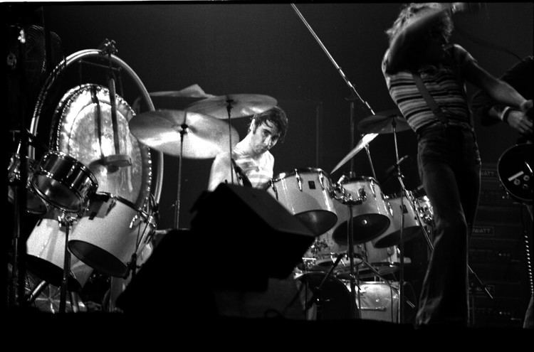 Keith Moon playing drums with two men on the right side singing and playing guitar at the Maple Leaf Gardens, Toronto. Keith with a serious face is wearing a striped shirt while the man on the right is wearing a striped shirt and pants