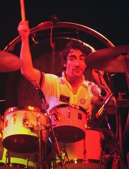 Keith Moon playing drums and looking at something with a serious face, mustache, and beard while wearing a white, black, and red polo shirt