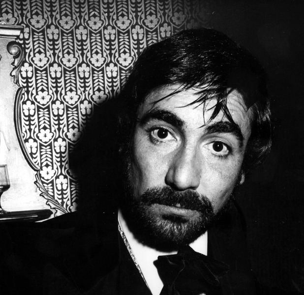 Keith Moon with a serious face, mustache, and beard while wearing a long sleeve under a bow tie and coat