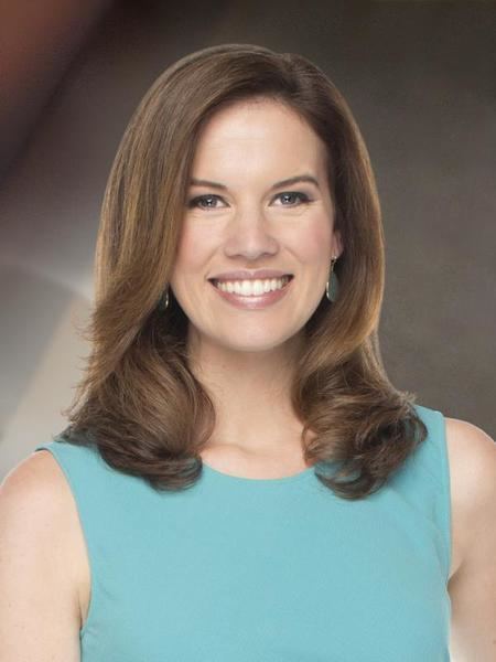 Kelly Evans smiling, with wavy hair, wearing earrings and a teal sleeveless dress.