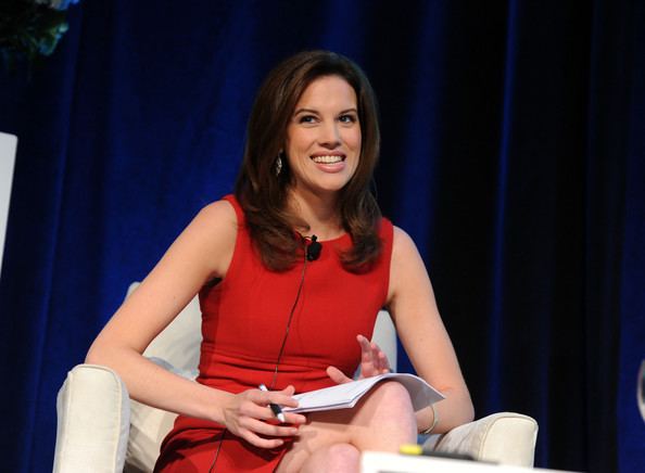 Kelly Evans smiling while sitting on a white couch and holding a paper and pen, with wavy hair, and wearing a red sleeveless dress.