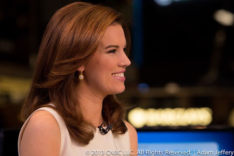 Kelly Evans side-view while smiling, with wavy blonde hair, wearing earrings, and a white sleeveless top.