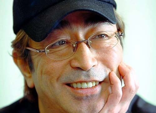 Ken Shimura smiling while hand on his chin and wearing a black cap, black shirt, and eyeglasses