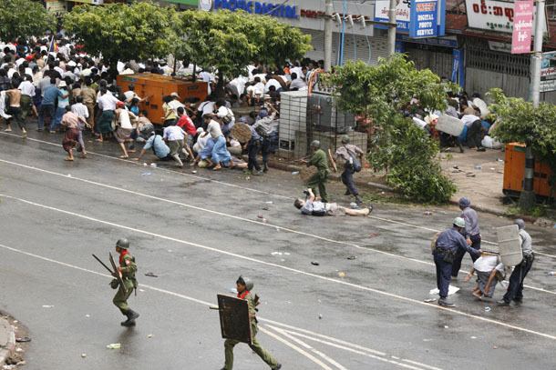 A street with a riot, at the right is a crowd running away from the police, trees soldiers in the middle wearing a dark green helmet, green uniform and slippers. in the ground, Kenji Nagai is laying taking photos of the riot, has black hair wearing a light blue polo, gray shorts and slippers. At the right is a police man wearing gray uniforms capturing a man sitting down, and a tow police officer has a stick, and riot shields, wearing a green uniform and green helmet.
