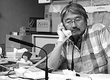 In black and white office, with a telephone on the table, Kenji Nagai is serious, sitting with his right arm resting at the table while holding a telephone, has black hair mustache, wears eyeglasses, and a checkered polo shirt.