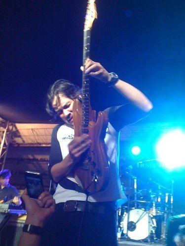 Kenneth Ilagan is serious, performing at a concert event using his brown guitar fretboard up, on top of a stage with blue lights and drums set at the back, he has black wavy hair wearing a black watch, a white shirt with a black mid-length sleeve, a black belt with black pants. In front is a hand wearing a black strap watch holding a phone recording Kenneth’s performance.