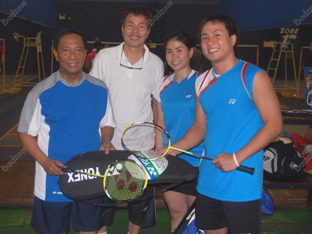 Inside a badminton court with with a yellow referee sits at the back, from left Jejomar Binay is smiling, standing holding a badminton racket and its bag on his left hand, he has black hair wearing a blue shirt with a gray shade across the shoulder and white at the side along with a black shorts, 2nd from left, Nelson Asucion is smiling standing at the back has black hair and a mustache wearing a white polo shirt, black shorts and a eyeglasses on his neck, 3rd from left, Kennie is smiling, standing at the back she has black hair wearing a blue badminton uniform, and black shorts. At the right, Kennevic Asuncion is smiling, standing, with his right hand holding a racket bag, and left holding a badminton racket, he has black hair wearing a blue badminton uniform with black shorts, and a white bracelet.